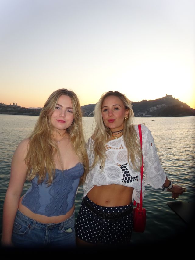 Dos chicas posan de pie frente al mar al atardecer, una con top azul de encaje y otra con blusa blanca, short de lunares y bolso rojo.