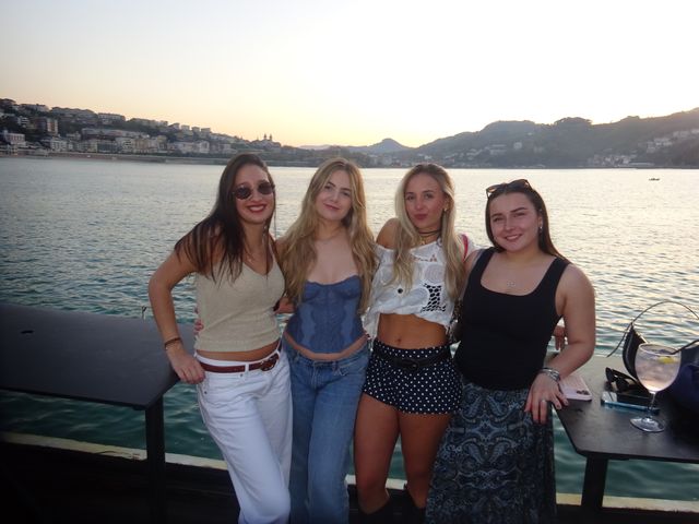 Cuatro amigas sonríen a la cámara de pie frente al mar al atardecer, una de ellas con gafas de sol redondas, junto a una mesa con una copa.