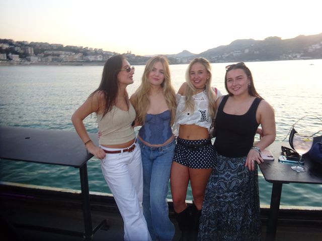 Un grupo de cuatro chicas posa sonriendo junto al mar al atardecer, con la ciudad a lo lejos y una copa sobre la mesa de detrás.