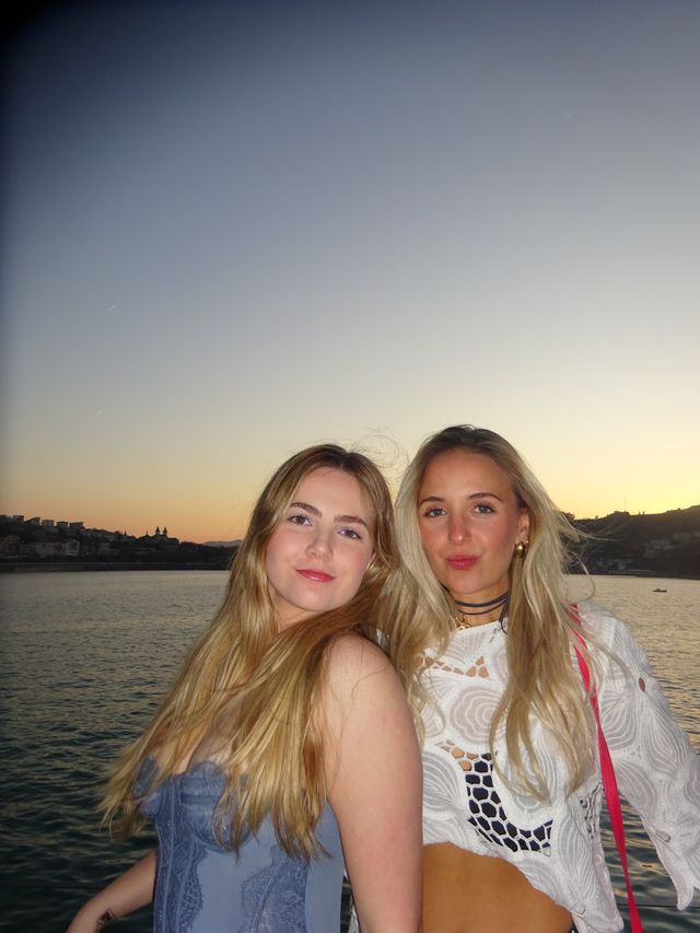 Dos chicas de pelo largo posan con las cabezas juntas y miran a la cámara, con el cielo del atardecer y el mar detrás.
