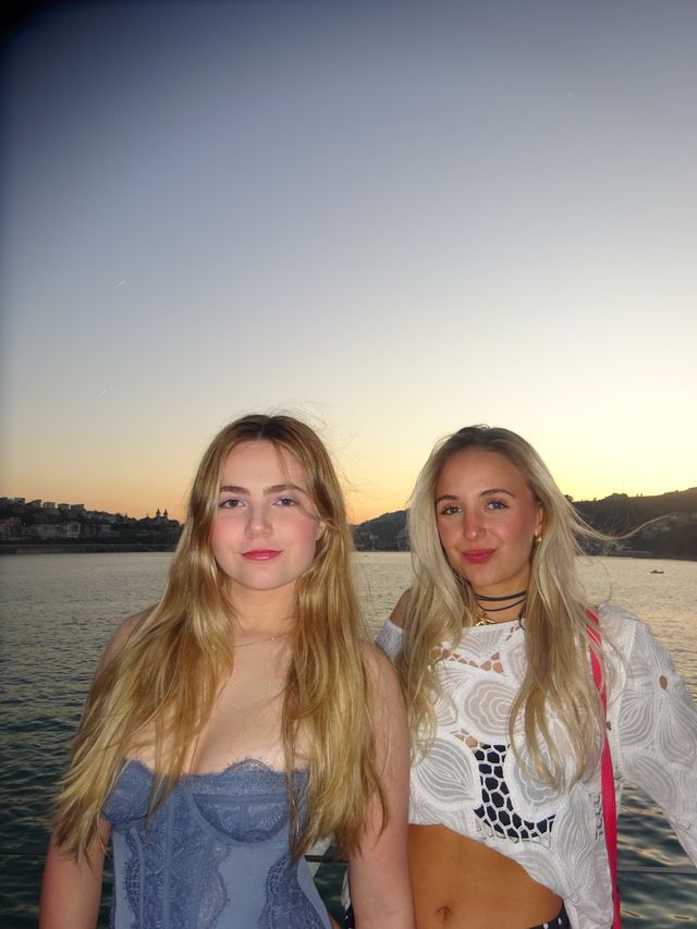 Dos chicas posan muy juntas y sonríen levemente a la cámara, una con top azul de encaje y otra con blusa blanca, contra un cielo claro.