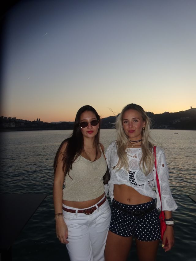 Dos chicas posan de pie junto al mar al atardecer, una con blusa blanca y short de lunares y otra con gafas de sol y pantalón blanco.