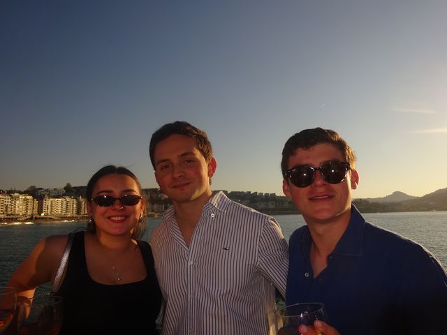 Una chica y dos chicos posan juntos y sonríen a cámara bajo la luz dorada del atardecer, con el mar y la bahía detrás.