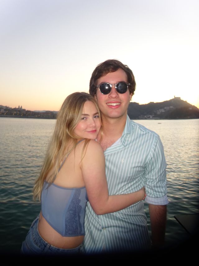 Una chica y un chico se abrazan sonrientes junto al mar al atardecer, él con gafas de sol redondas y camisa de rayas verdes.