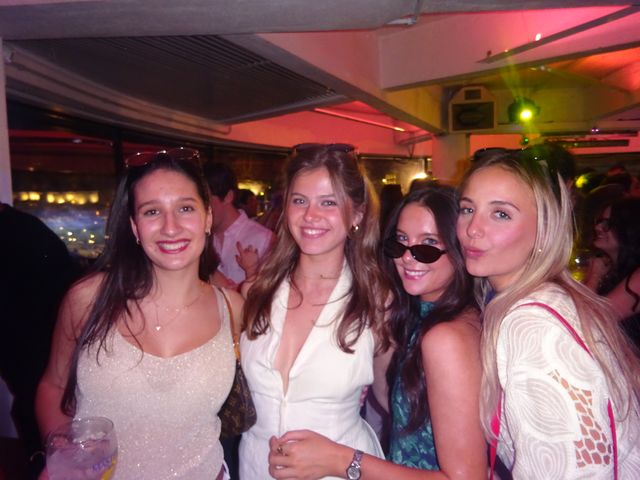 Cuatro chicas posan muy juntas, tres sonríen y una hace morritos, en un local con luz rosada y un foco verde; la de la izquierda sostiene un vaso.