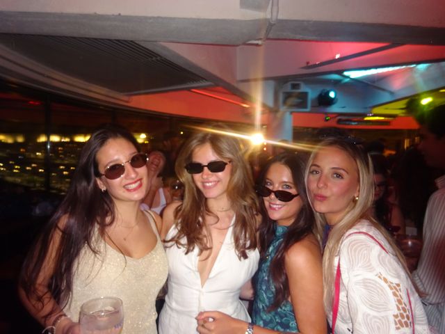 Cuatro chicas posan juntas bajo luces rojas, tres con gafas de sol y otra haciendo morritos, con un destello de luz al fondo.