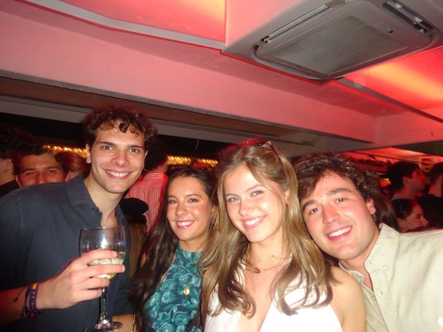 Dos chicos y dos chicas posan muy juntos y sonríen a cámara bajo un techo iluminado de rosa; uno de ellos sostiene una copa.