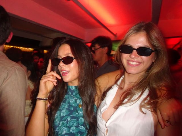 Dos chicas con gafas de sol posan abrazadas bajo una luz roja; una levanta el dedo mientras habla y la otra sonríe a cámara.