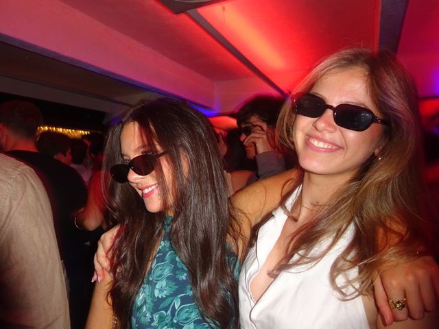 Dos chicas con gafas de sol posan abrazadas entre luces rojas y moradas; una sonríe mirando hacia abajo y la otra, a cámara.