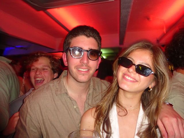 Un chico con gafas y una chica con gafas de sol posan abrazados y sonrientes bajo un techo rojo; detrás, otro chico se ríe a carcajadas.