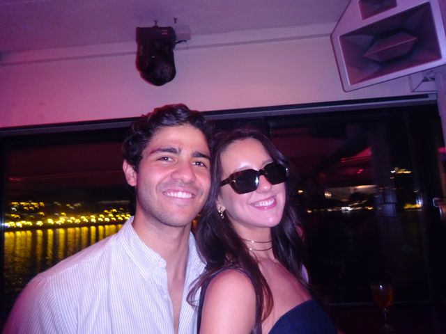 Una chica con gafas de sol y un chico posan sonrientes junto a una ventana, con luces amarillas reflejadas en el agua al fondo.