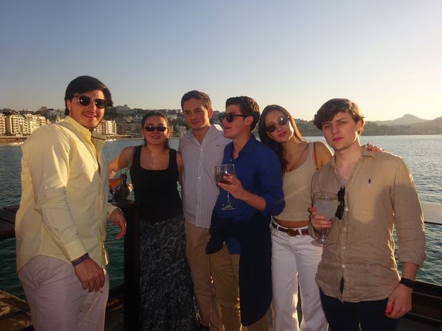 Un grupo de dos chicas y cuatro chicos posa junto al mar con copas en la mano, con la bahía iluminada por el sol del atardecer.