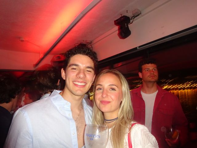 Un chico y una chica rubia posan sonrientes bajo una luz roja, ella con una copa grande; detrás, un chico con chaqueta roja mira a otro lado.