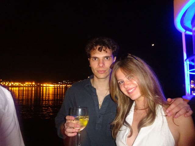 Un chico con una copa y una chica sonriente posan abrazados de noche frente al agua, con luces de ciudad y un neón azul detrás.
