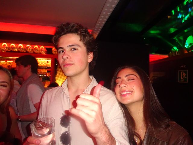 Un chico con camisa clara levanta el pulgar y una chica sonríe con los ojos cerrados a su lado, ante una barra con luz roja.