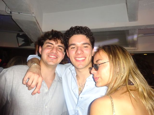 Dos chicos de pelo rizado se abrazan sonrientes junto a una chica rubia con gafas de sol, bajo un techo gris con tuberías.