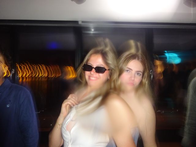 Dos chicas posan juntas con un efecto de movimiento, una sonríe con gafas de sol oscuras y detrás se ven trazos de luz naranja.