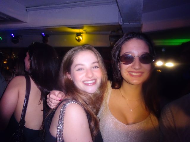 Dos chicas sonríen a la cámara abrazadas, una con gafas de sol ovaladas, bajo un techo con focos de luz amarilla, azul y verde.