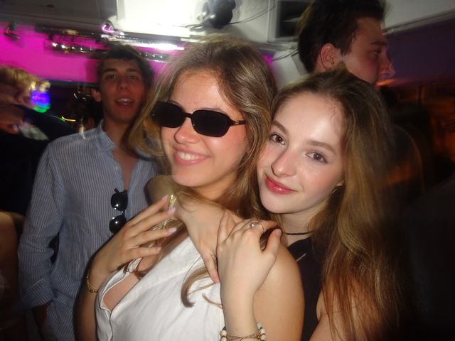 Dos chicas posan con las cabezas juntas, una con gafas de sol y otra de labios rojos, con chicos detrás y luces rosas al fondo.