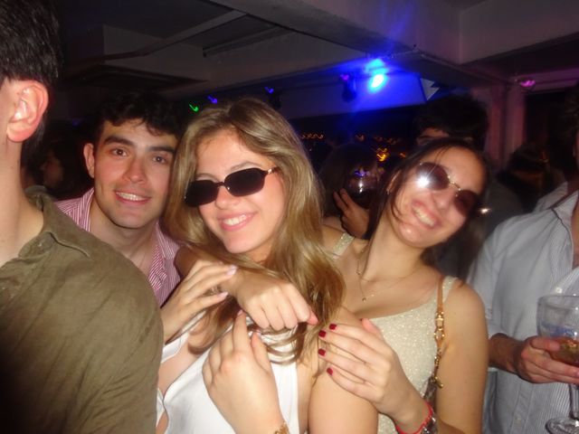 Dos chicas con gafas de sol sonríen abrazadas entre la gente, con un chico sonriente detrás y luces azules y moradas en el techo.