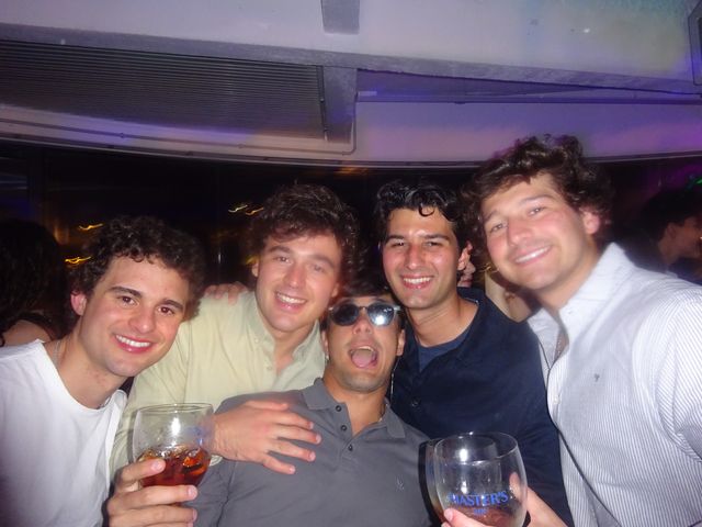 Cinco chicos posan apretados y sonrientes, dos con copas en la mano y uno con gafas de sol y la boca abierta, bajo luz morada.