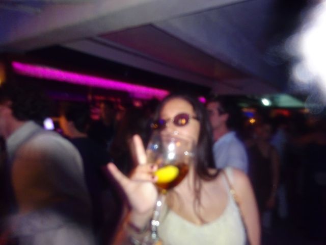 Una chica con gafas de sol sostiene una copa con limón y hace un gesto con la mano, en un local con luces rosas y mucha gente detrás.