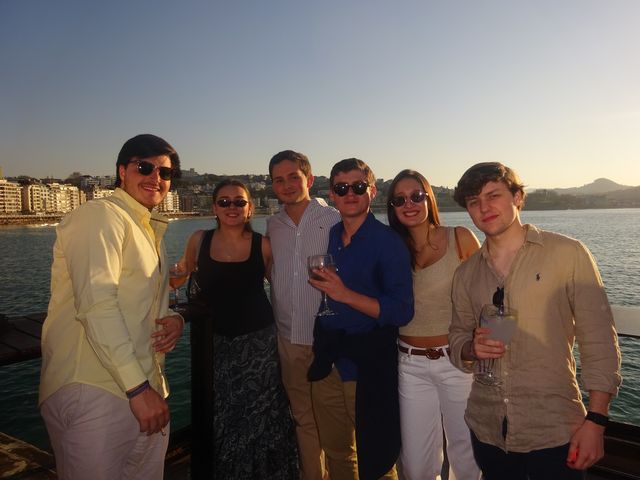 Dos chicas y cuatro chicos sonríen a cámara agrupados junto a una barandilla, con el mar y la bahía detrás al atardecer.