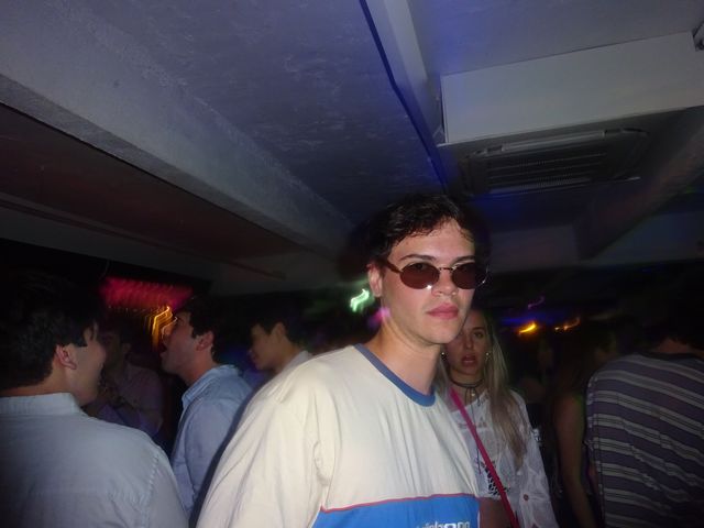 Un chico con gafas de sol y camiseta blanca con detalles azules mira serio a la cámara entre la gente, en un local oscuro de techo bajo.