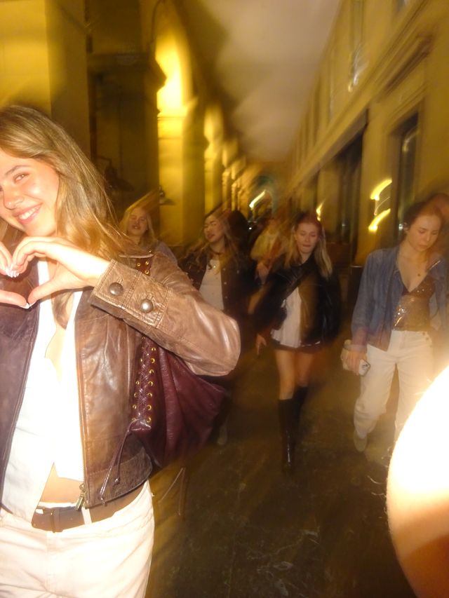 Una chica sonríe y forma un corazón con las manos mientras un grupo de amigas camina detrás por unos soportales iluminados de noche.