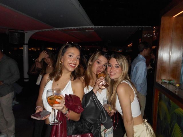 Tres chicas posan juntas con copas en la mano en un bar oscuro de techo rojo, con más gente al fondo.