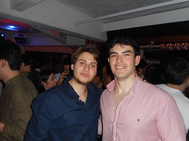 Dos chicos posan juntos y sonríen a la cámara en un local lleno de gente, uno con camisa azul marino y otro con camisa de rayas rosas.