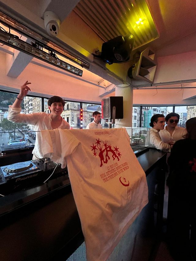 Un DJ con camisa blanca sonríe y levanta la mano detrás de los platos, mientras una camiseta blanca de estampado rojo cuelga de la cabina.