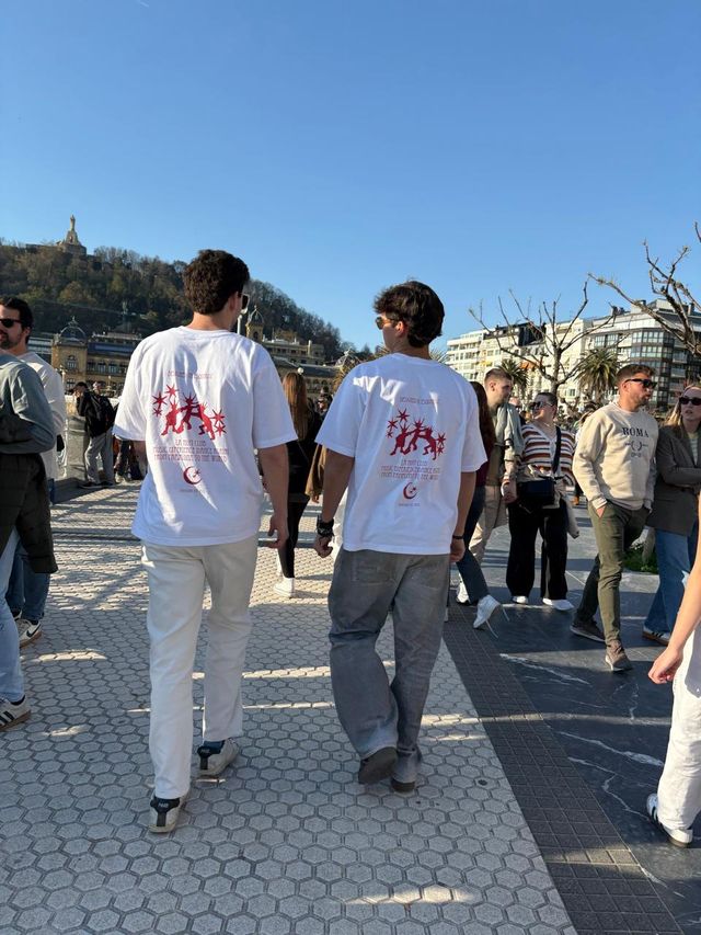 Dos chicos con camisetas blancas de estampado rojo caminan de espaldas por una acera soleada de baldosas hexagonales, con una colina al fondo.