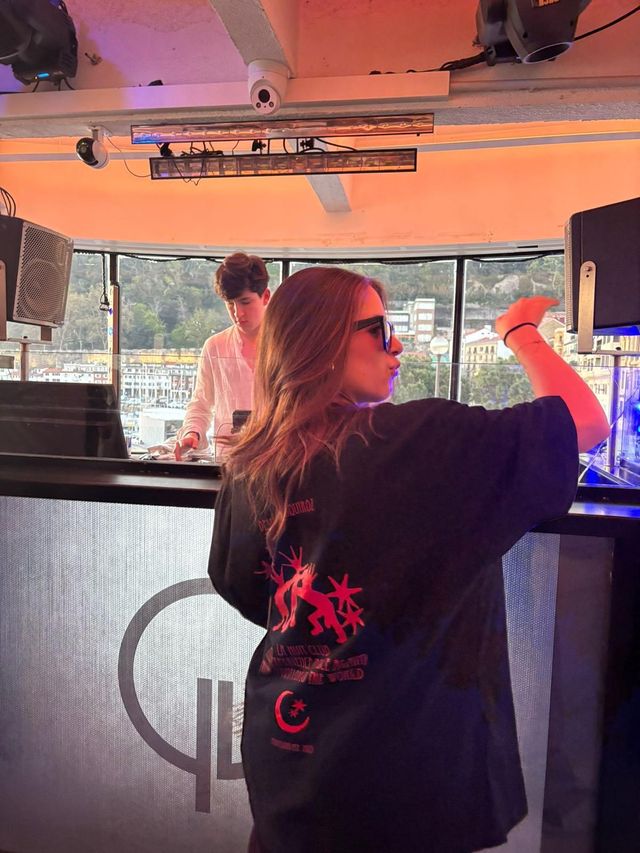 Una chica con gafas de sol y camiseta negra de estampado rojo baila con un brazo en alto, mientras un DJ de camisa blanca pincha detrás.