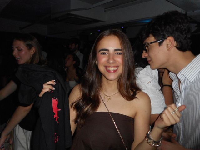 Una chica sonríe a la cámara en una sala oscura mientras un chico con gafas y camisa de rayas mira hacia ella, rodeados de gente.
