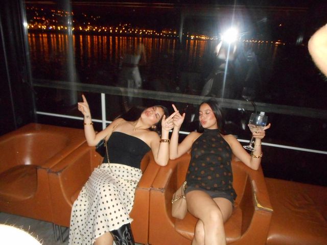 Dos chicas posan reclinadas en sillones naranjas, con los ojos cerrados y los dedos en alto, junto a un ventanal con luces sobre el agua.