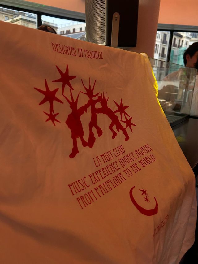 Primer plano de una camiseta blanca con estampado rojo de figuras, estrellas y el texto La Nuit Club, colgada junto a un ventanal.