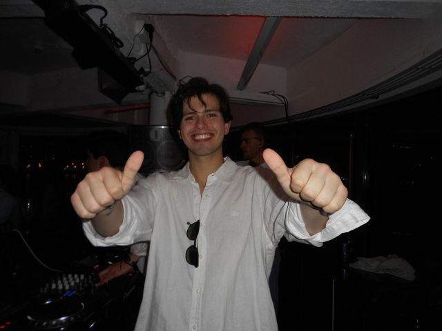 Un chico con camisa blanca sonríe y levanta los pulgares a la cámara junto al equipo de DJ, en una sala oscura.
