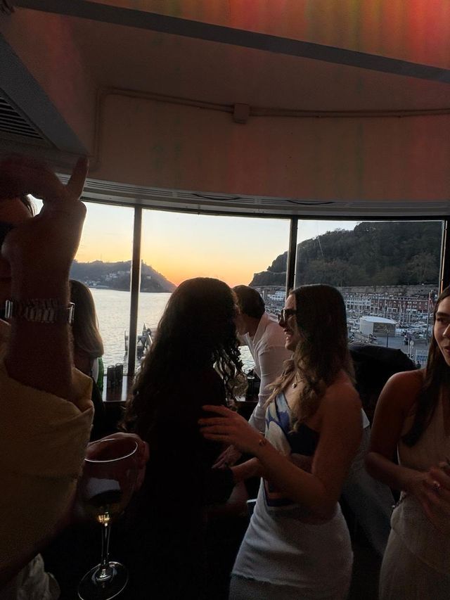 Un grupo de chicas y chicos charla a contraluz ante un gran ventanal, con el atardecer naranja sobre la bahía y una copa de vino delante.
