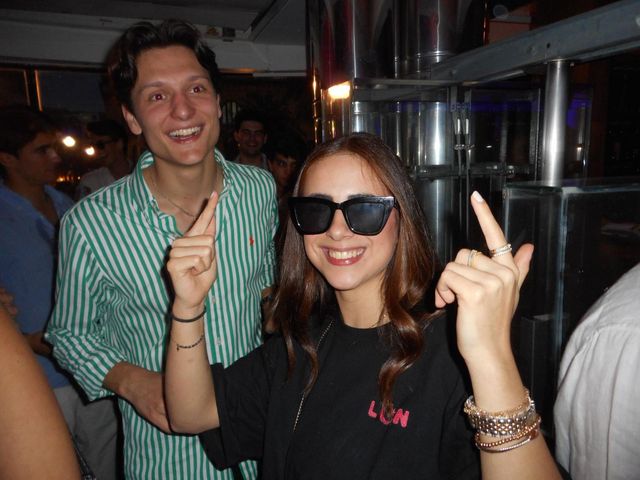 Una chica con gafas de sol y un chico con camisa de rayas verdes sonríen, y ella señala hacia arriba con las dos manos, ante cilindros metálicos.