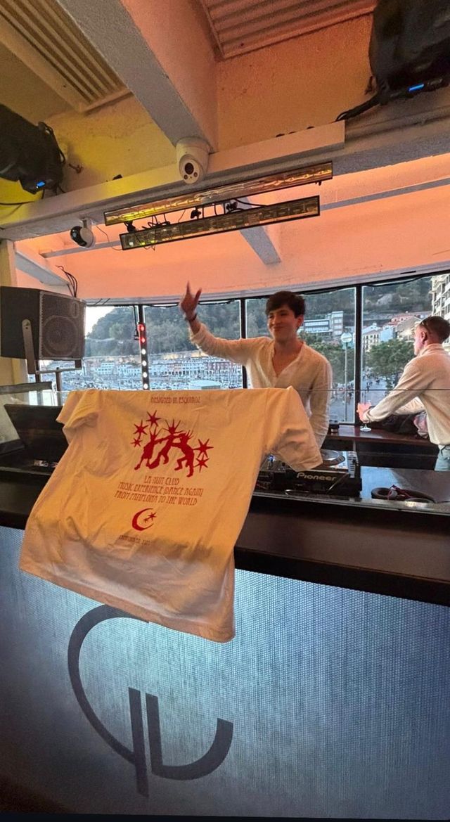 Un DJ con camisa clara sonríe con el brazo en alto tras los platos, con una camiseta de La Nuit Club colgada de la cabina y el puerto detrás.