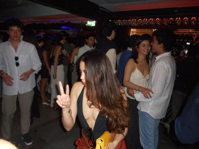Una chica con top negro hace el signo de la paz en una sala llena de gente, con una pareja abrazada detrás y la barra iluminada al fondo.