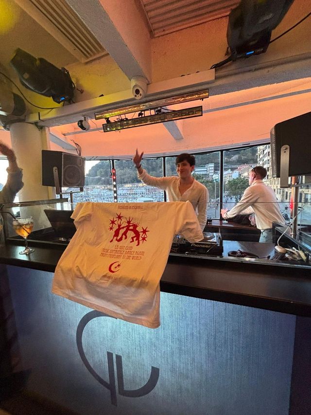 Un DJ sonríe con el brazo en alto tras los platos, junto a un chico con una copa, y delante cuelga una camiseta con estampado rojo.