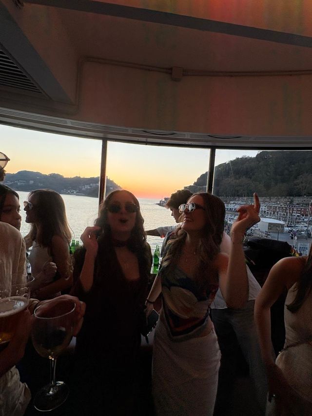 Un grupo de chicas con gafas de sol baila junto al ventanal, una con la mano en alto, y el cielo naranja del atardecer al fondo.