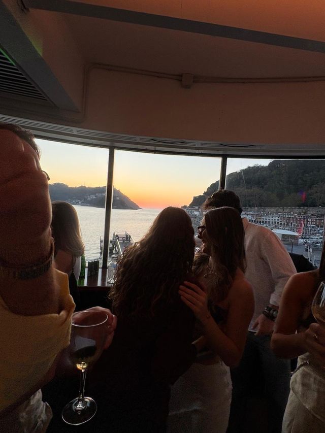 Dos chicas charlan muy juntas ante el ventanal, una de espaldas, con un chico de camisa blanca detrás y el atardecer sobre la bahía.