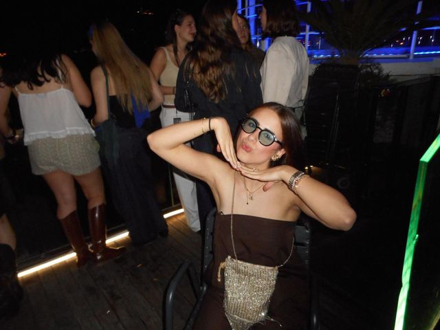 Una chica sentada con gafas redondas y top marrón posa con las manos bajo la barbilla en una terraza de noche, con gente de pie detrás.