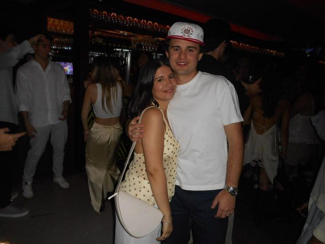Una pareja sonriente posa abrazada en una sala oscura, él con gorra blanca y ella con top de lunares, y detrás se ve gente y la barra.