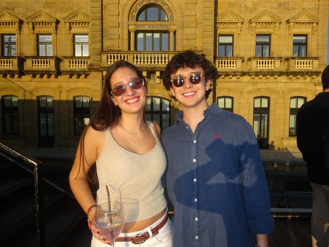 Una chica y un chico con gafas de sol posan sonrientes frente a un edificio señorial de piedra dorada bañado por el sol.