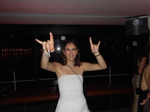 Una chica con vestido blanco sin tirantes sonríe y levanta las dos manos con el gesto del rock, con un ventanal oscuro y luces lejanas detrás.