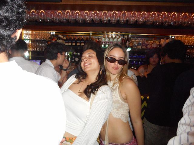 Dos chicas posan juntas delante de la barra de un bar, una sonríe con los ojos cerrados y la otra lleva gafas de sol; detrás, copas y botellas.